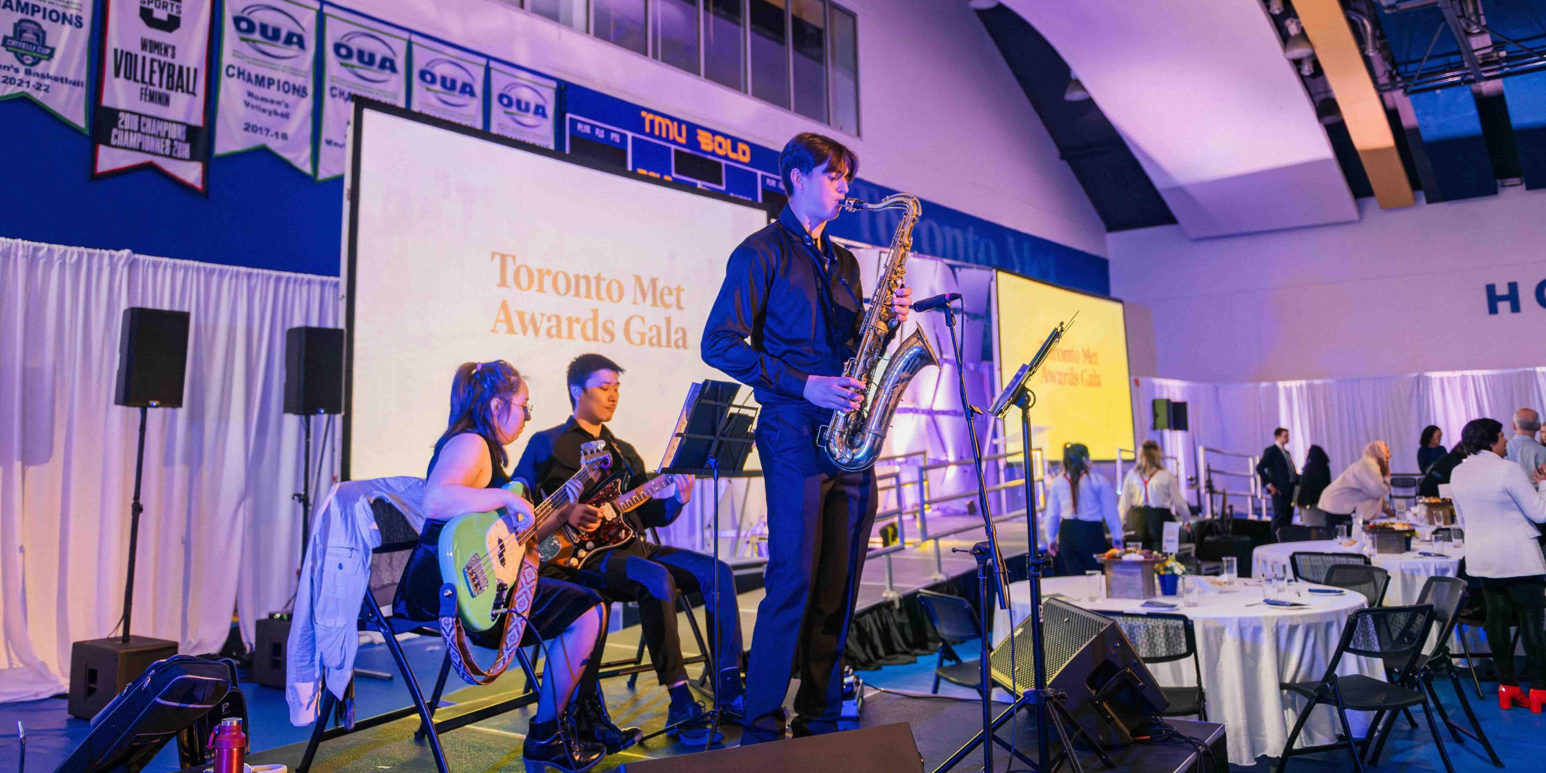 A band of three performing at the Toronto Met Awards Gala. Two are seated, one is standing playing the saxophone.